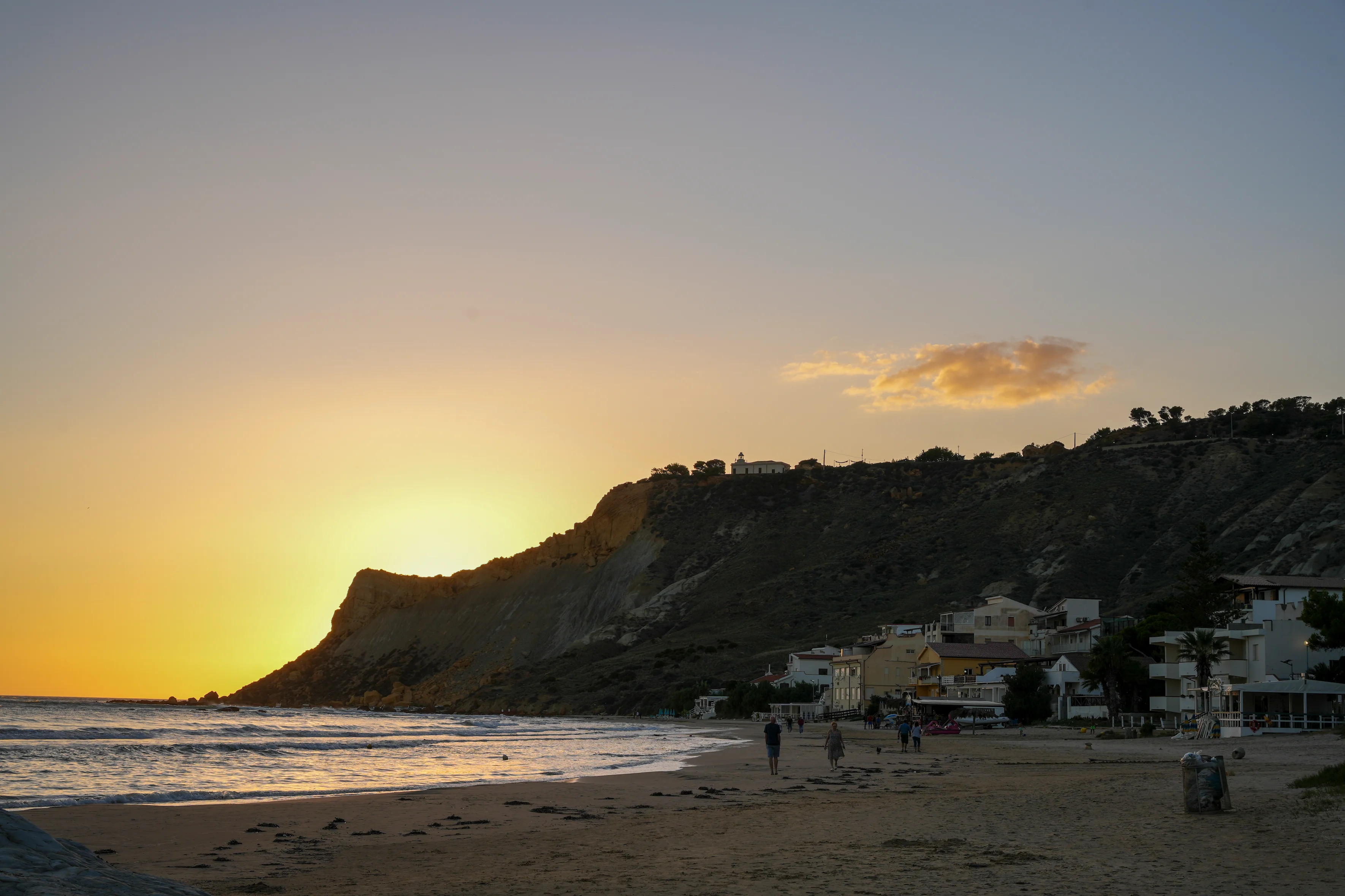 Sunset over a beach with gentle waves, silhouetted cliff, and houses along the shore.