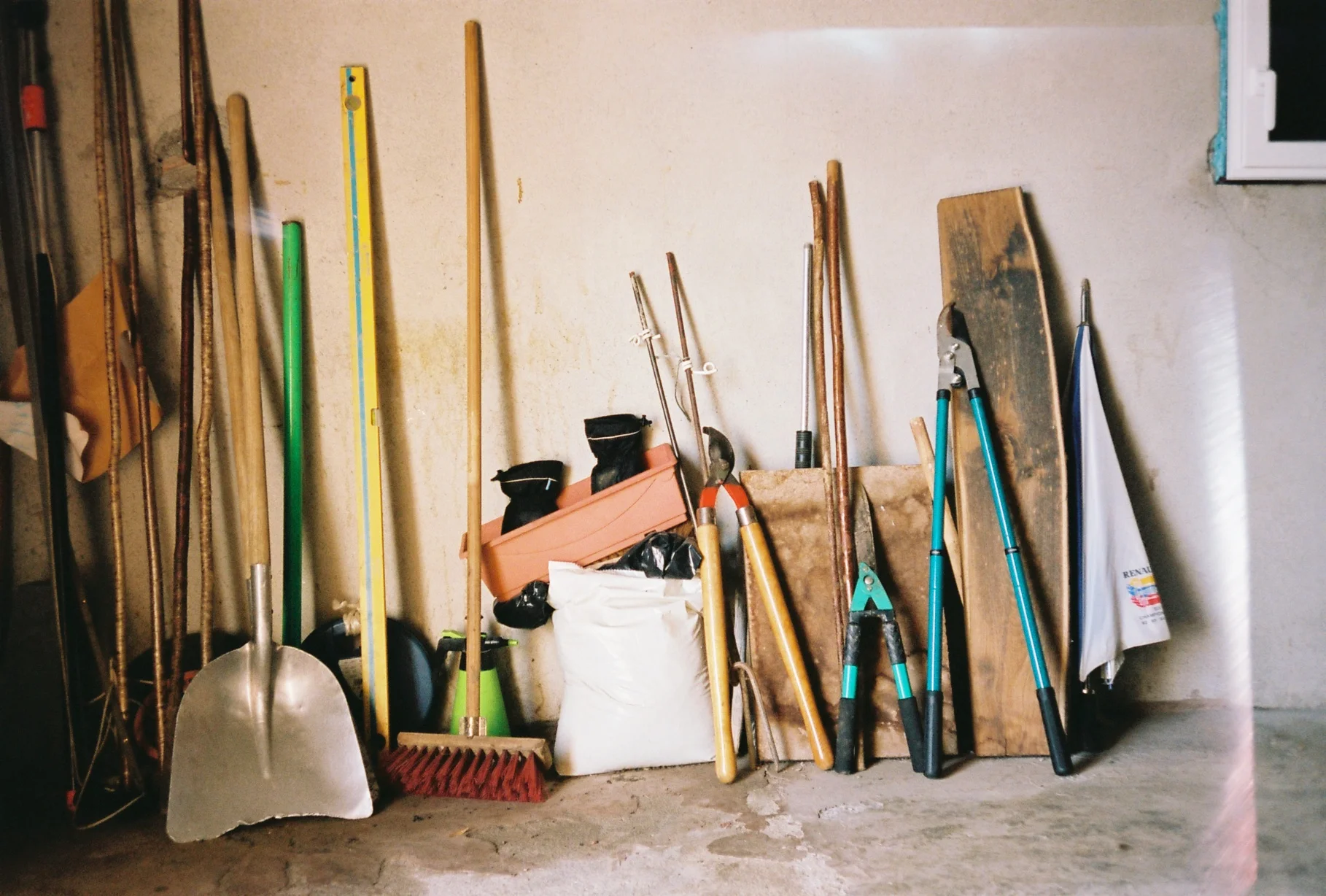 Various gardening and cleaning tools including a shovel, broom, shears, and wooden sticks, leaning against a wall in a storage room.