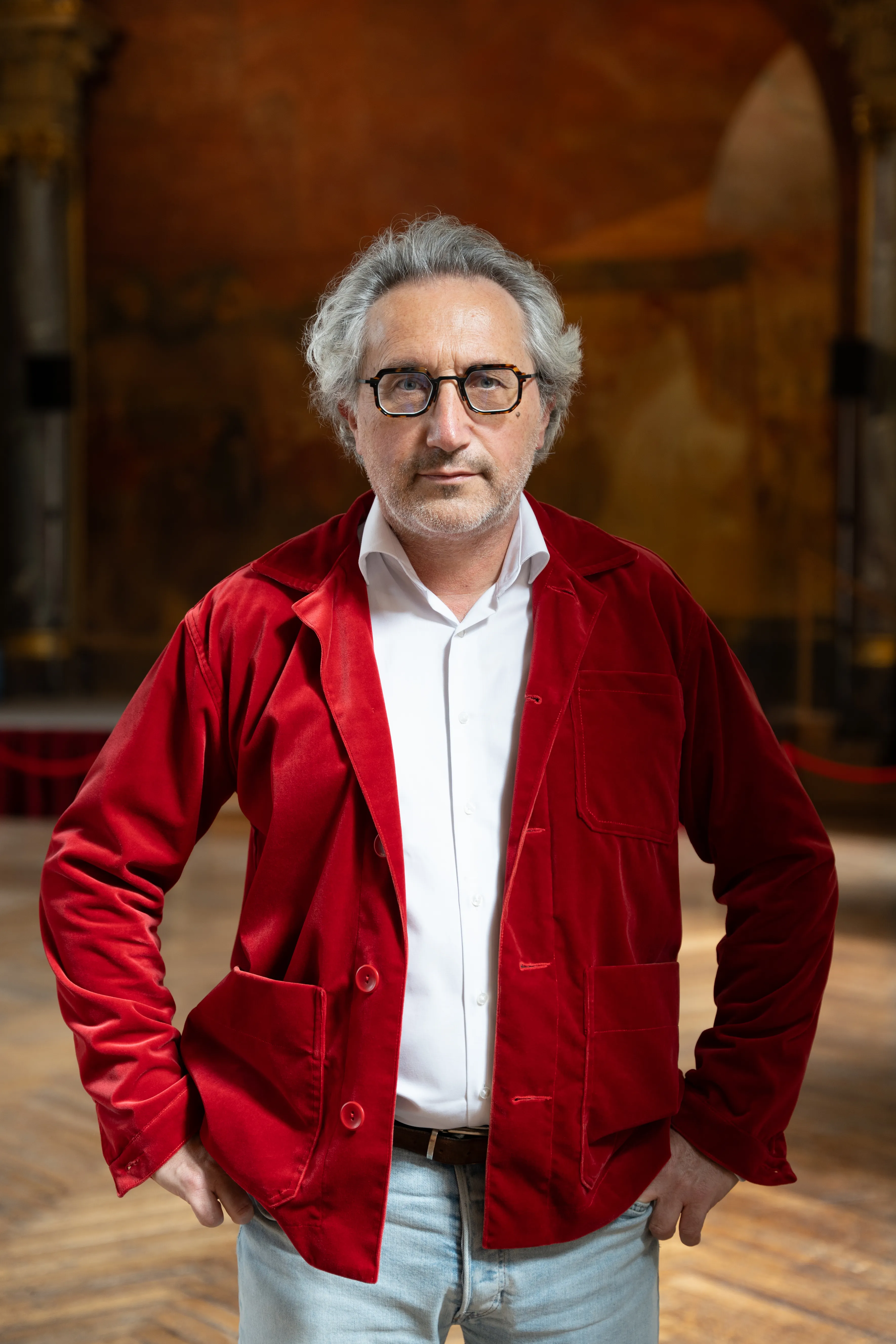 Middle-aged man with gray hair and glasses wearing a red velvet jacket over a white shirt, standing indoors with hands on hips.