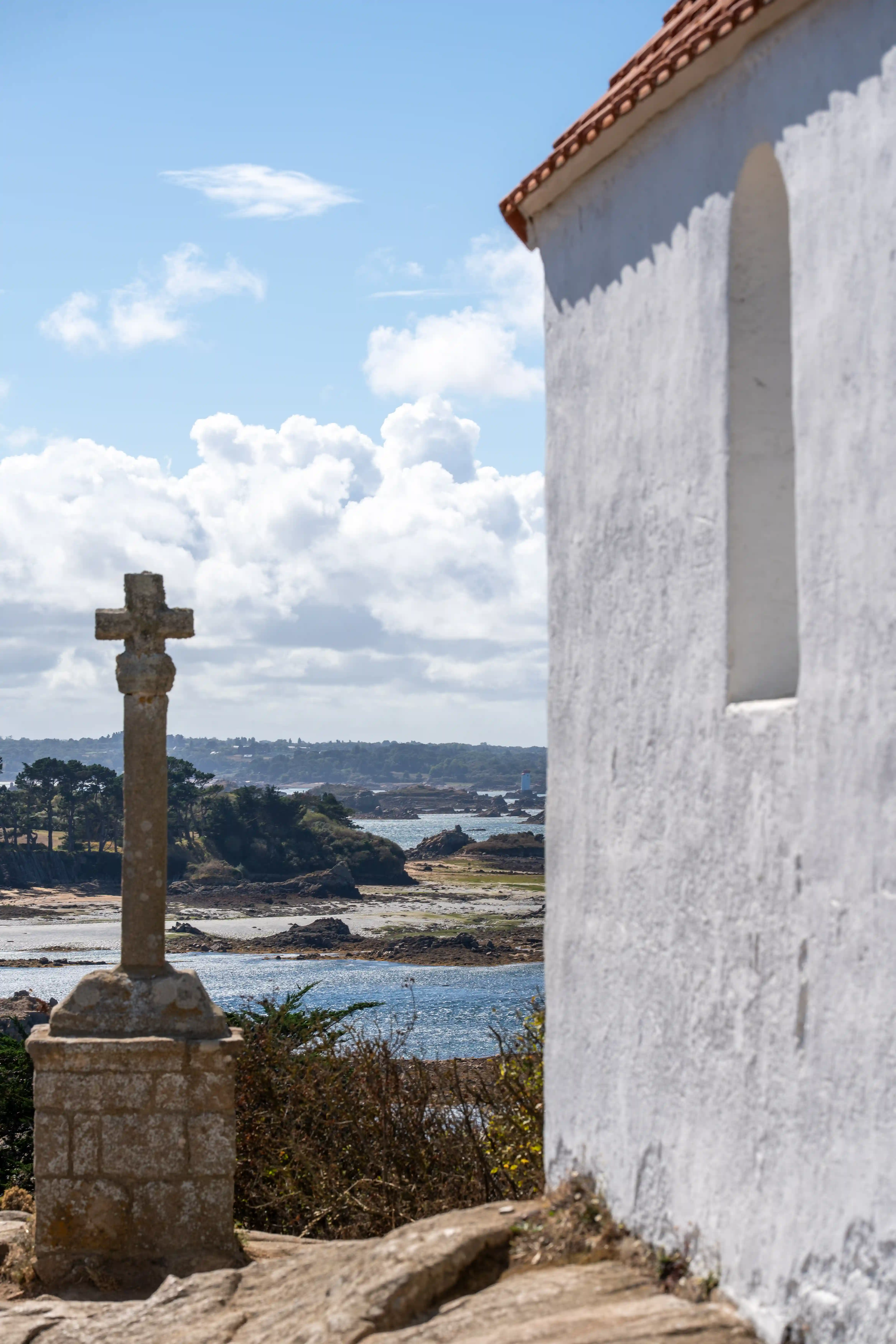 Stone cross monument near a white building overlooking a rocky coastline and blue water under a partly cloudy sky.