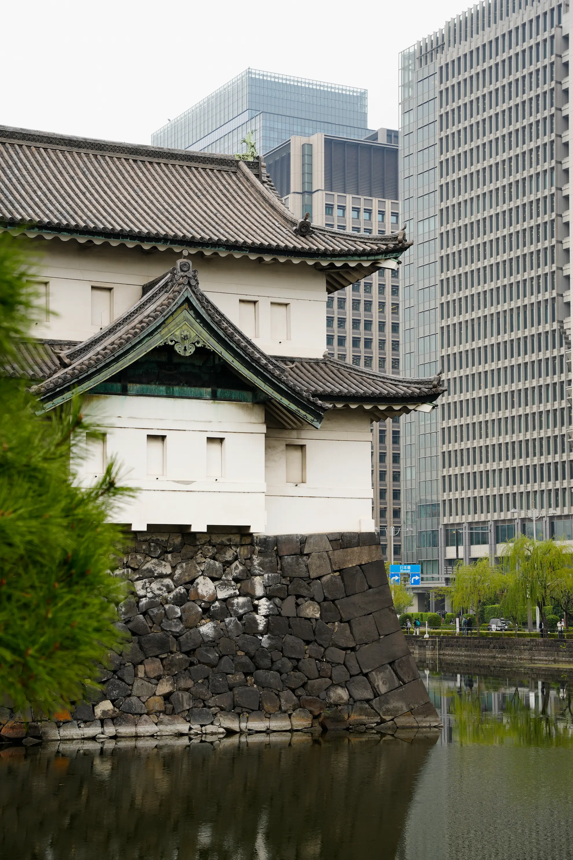 Traditional Japanese castle corner with tiled roof and stone foundation beside a moat, set against modern skyscrapers.