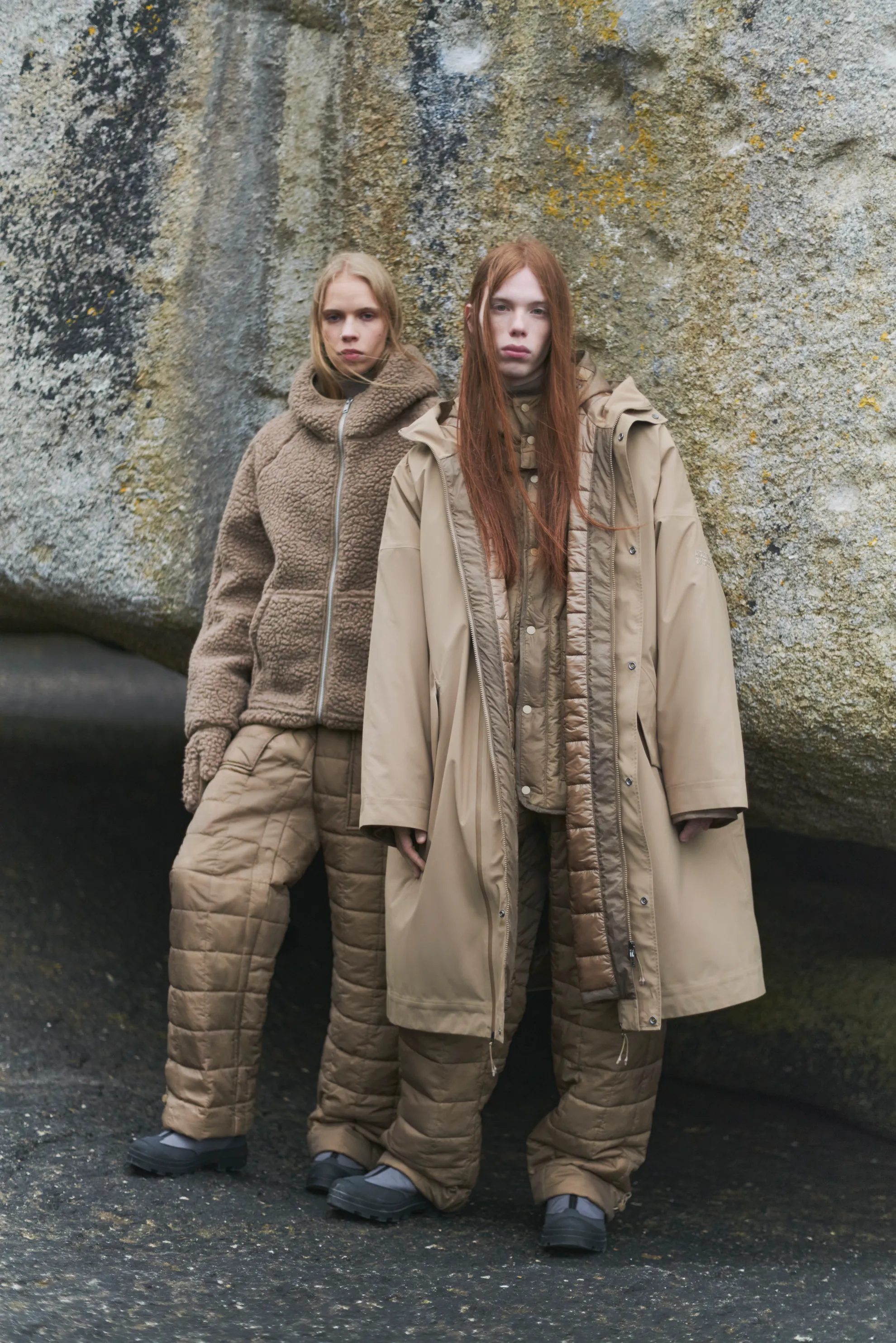 Two women standing outdoors in front of a large rock wearing layered neutral-toned winter clothing with quilted pants and jackets.