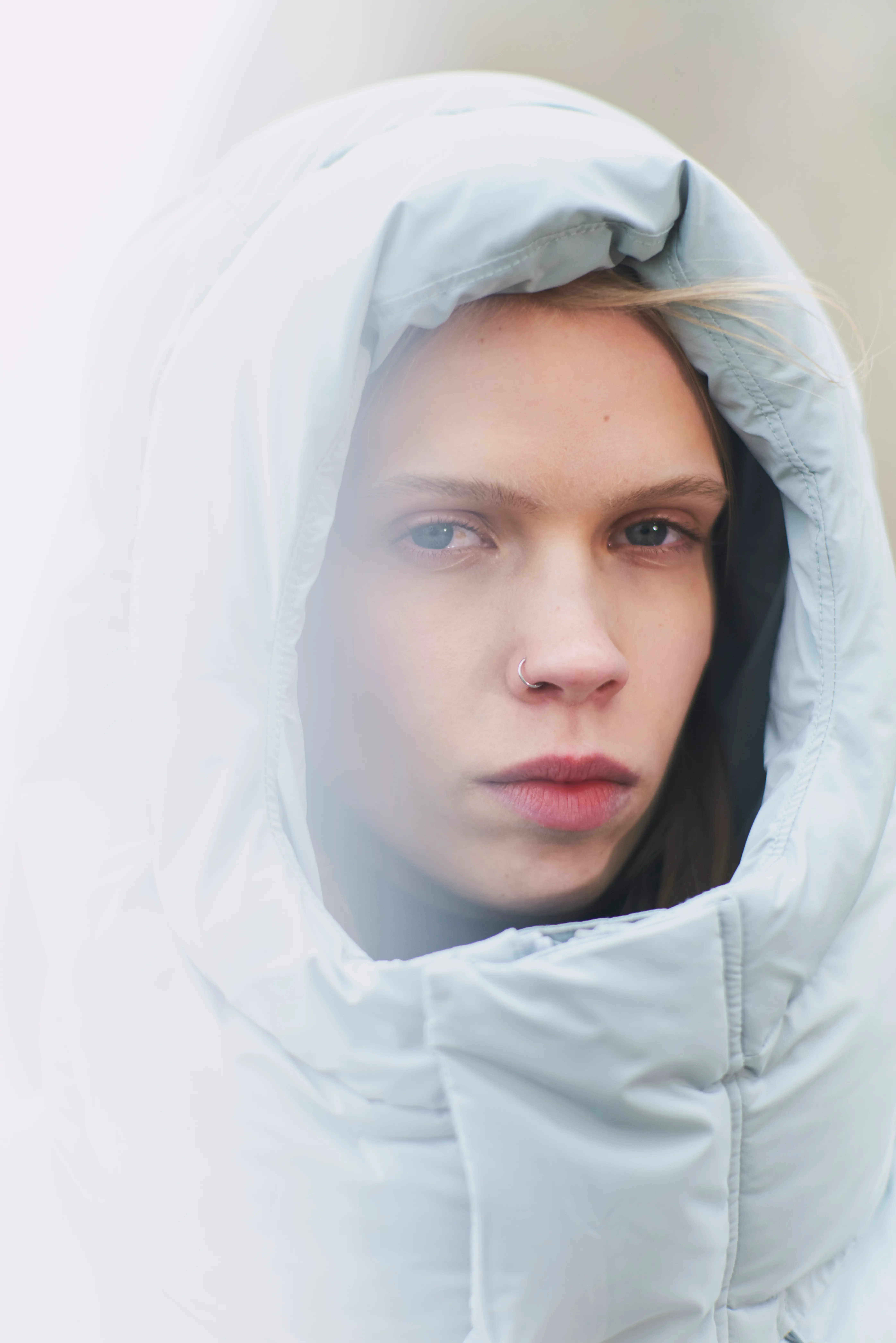 Close-up of a person with light blue eyes wearing a large light gray hooded jacket and a nose ring.